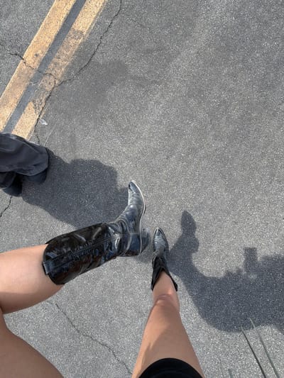 The image shows two people standing on asphalt. One person is wearing a shiny, black, knee-high boot on their right leg and gray pants. The other person is wearing a black shoe. The location is likely a street or parking lot in Palm Desert, California, United States of America. Evidence of this assumption includes asphalt ground that is typical of many streets and parking lots, and the asphalt having yellow pavement lines. There is a shadow that indicates the time of day is during daylight hours.
