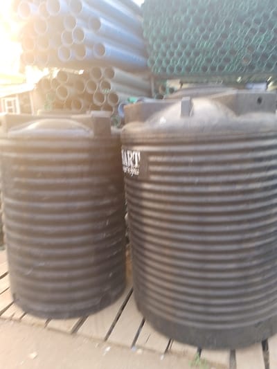 The image displays an outdoor storage or sales area for plumbing and construction materials, illuminated by bright daylight. In the foreground, two large, black, cylindrical water storage tanks are prominently featured. These tanks are made of plastic or a similar material and have a distinct ribbed or corrugated exterior. The tank on the right has a white label on its side, clearly showing the brand name "SMART" in bold capital letters, with smaller, illegible text below it, likely detailing the product or manufacturer. Both tanks are resting on wooden pallets or planks.
Behind the tanks, numerous pipes are stacked. To the left and top-center, large quantities of light grey or white PVC pipes are arranged horizontally. To the right, another stack of green pipes, possibly also PVC, is visible. The intense sunlight creates a significant glare or lens flare in the upper left corner of the image, indicating a clear, sunny day. There are no people visible, and no specific activity is taking place; the scene depicts an inventory of goods. Given the context, this scene is located in Ijaiye, Nigeria, showcasing common supplies for water infrastructure or building.