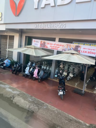 A storefront reads "XE ĐIỆN LÂM HỒNG" in large letters. Banners advertise "CÙNG VADEA - BỐC THĂM LIỀN TAY" and "KHU VỰC BẢO HÀNH CÔNG TY TNHH XE MÁY LÂM HỒNG" alongside YADEA and AQUA logos. Numerous scooters are parked outside under large sun umbrellas. A motorcycle license plate is visible with the numbers "47-B2 202.02". The scene is captured from a slightly angled perspective, with the edge of a street or sidewalk visible on the right.