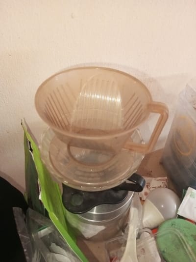 An elevated, transparent coffee filter rests on a matching base above a metal coffee pot. The filter is tinted a light tan and features a distinct handle. A green and white package stands beside the pot. The surrounding area is cluttered with miscellaneous items including a lightbulb, disposable spoon, and what appears to be a lid, all on a surface against an off-white wall.