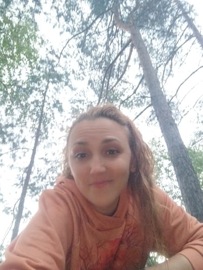 A selfie of a woman taken from a low angle, looking up at the camera. She has light brown hair and is wearing an orange hooded sweatshirt. The background is a canopy of trees against a bright sky, suggesting an outdoor setting. The location is specified as Tarasovskoe, Russia.