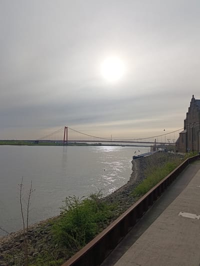 The image shows a view of Emmerich on the Rhine, Germany, featuring the Emmerich Rhine Bridge (Rheinbrücke Emmerich). The red suspension bridge spans the Rhine River, connecting Emmerich with Kleve. The sun is obscured by cloud cover, creating a diffused light across the sky. The Rhine River is visible, reflecting the sunlight. On the right side of the frame is a section of the city, including a brick building with architectural details, likely a church. A concrete walkway runs along the riverbank, separated from the water by a low, rusted metal barrier and a stone embankment. Sparse vegetation is present along the riverbank. Several people can be seen walking along the walkway. A barge is docked along the shoreline. A bird is visible in flight near the building on the right side.