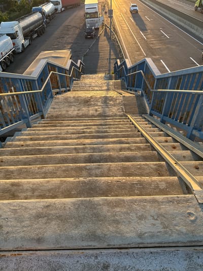 A concrete pedestrian overpass with weathered blue metal railings spans over a highway. The overpass descends towards the viewer, with rows of concrete steps clearly visible. The highway below is paved and marked with white dashed lines, with traffic visible in the distance. On the left side of the overpass, parked semi-trucks, including tanker trucks, are lined up on a shoulder or service area. A small dark car is visible in the distance on the right side of the highway. The scene is illuminated by the warm, low light of the setting sun, casting long shadows.