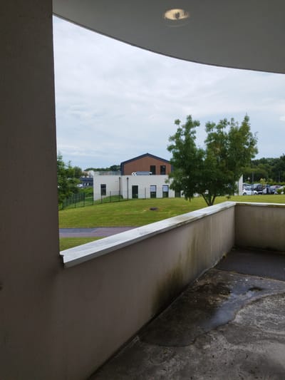 This media file captures an outdoor scene, likely from a balcony or elevated vantage point, looking out onto a modern building complex and its surroundings. The foreground features a textured concrete wall and a light-colored railing, showing signs of weathering and some water stains on the ground below, suggesting recent rain or a damp environment.
Beyond the railing, a well-maintained lawn slopes gently towards a low fence. In the mid-ground, a modern, light-colored building with dark-trimmed windows and a distinctive brown, angled roof dominates the landscape. Adjacent to it is another building, also white with dark windows. A lone, mature tree with lush green foliage stands prominently in front of the buildings, adding a natural element to the architectural scene.
In the background, a parking lot is visible with several cars, including a white one, and a dense line of trees forming the horizon. The sky is overcast with soft, diffused light, indicating a cloudy day, possibly late afternoon or early morning given the muted light. The location is identified as Cherbourg-en-Cotentin, France. There is no visible text within the image. The overall mood is calm and somewhat subdued, reflective of the overcast weather.