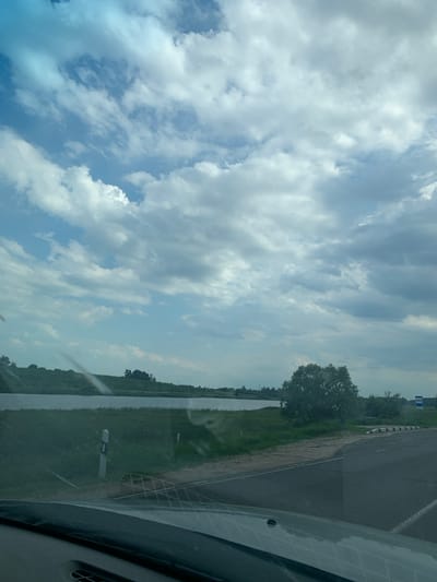 The image depicts a roadside view on a cloudy day. A paved road curves to the right, with a grassy embankment and a body of water on the left. Several trees are visible on the far bank of the water and further inland. A road sign is present on the right side of the road. The foreground shows the dashboard and windshield of a vehicle, suggesting the photo was taken from a car. The sky is filled with dramatic clouds. The location is identified as Слабада-Кучынскі сельскі Савет, Belarus.