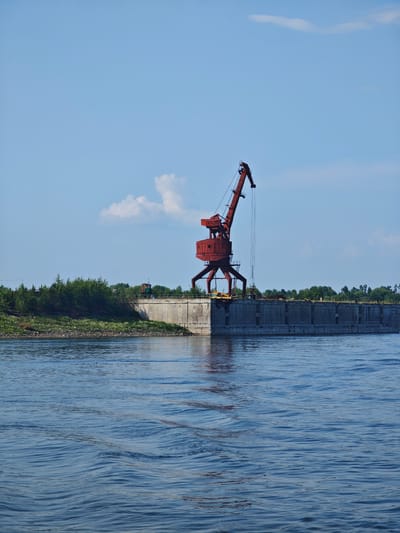 A red gantry crane stands on a concrete pier on the bank of a wide, blue river under a bright blue sky with a few scattered white clouds. The pier is lined with a concrete wall and a railing. Behind the pier, there is a line of green trees and bushes. The water in the foreground is rippled, suggesting a slight current or breeze. The scene appears to be an industrial waterfront, possibly a port or loading facility. There are no people visible in the image. The location is the city of Verkhnepashino, Russia.