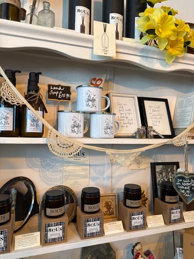 This image depicts a three-tiered white painted wooden shelving unit, likely inside a retail establishment in Ambleside, United Kingdom, displaying various home goods and decor items.

On the top shelf, from left to right, are two dark glass bottles, two clear glass bottles (one slender, one wider), and multiple black cylindrical boxes labeled "BOUS Eco Diffuser with Essential Oils." A white card with "Diffuser 150g £39" and a graphic is visible, along with a smaller card stating "Havana." To the far right, a bunch of artificial yellow flowers is positioned.

The middle shelf features two dark amber pump bottles, each labeled "Mr. H's Tearoom HAND SOAP" and adorned with twine. A small black chalkboard sign reads "Hand Soap £15.95." Three white enamel mugs with silver rims, depicting a pig on a bicycle and labeled "Mr. H's," are stacked to the right of the soap bottles. A white crocheted lace garland is draped across this shelf. To the right of the mugs are two framed pieces of art. The white-framed piece reads, "She's an old soul with young eyes, a vintage heart and a beautiful mind." The black-framed piece reads, "You Can't Start an adventure without a cup of tea." Two small glass vials with cork stoppers, one containing pink material and the other a small metal spout, are positioned in front of the frames. A large white sign describes "BOUS Natural Candles and Home Fragrance," mentioning it as a "small-batch candle brand" that is "Vegan" and has "No nasties." A smaller card states "100g Medium 25 hrs Burn time £16 Multi Sensory Wood Wick Candle." A partial view of a black-framed photograph is visible to the far right.

The bottom shelf displays multiple dark amber jars with black lids, labeled "BOUS," resting on small brown square risers. These appear to be candles, with specific scents such as "Ancient Woodland," "Havana," "Petrichor," "Rose Hammam," and "Lichen" indicated on labels below the jars. Each brown riser box is labeled "BOUS Aromatherapy Soy Candle Hand Poured Up North." A heart-shaped handheld mirror reflects a person holding a camera/phone. A round, ornate silver-framed mirror is also visible behind some products. A small vintage-style framed illustration is also present. To the far right, a heart-shaped metal plaque hangs, inscribed with text starting "you can't st..." and "an adventure without a cup of tea." Partially visible textile items are located at the bottom right.