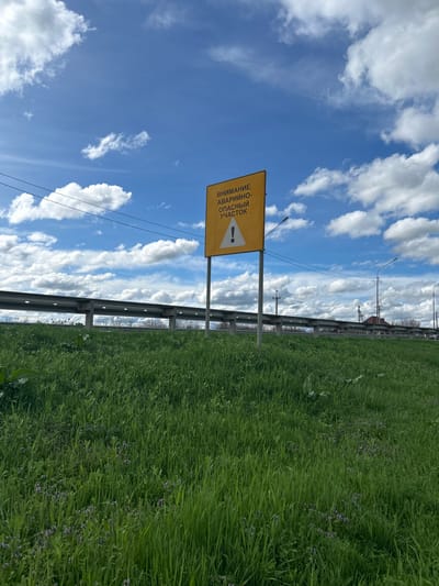 The image shows a roadside scene in Yugo-Severnaya, Russia. A yellow warning sign with the text "ВНИМАНИЕ АВАРИЙНЫЙ УЧАСТОК" (ATTENTION ACCIDENT-PRONE SECTION) and an exclamation point icon is visible next to a gray metal guardrail. Beyond the guardrail is a field of tall green grass with small purple flowers. Above the sign and grass is a blue sky with numerous white clouds. Power lines run parallel to the guardrail and the road.