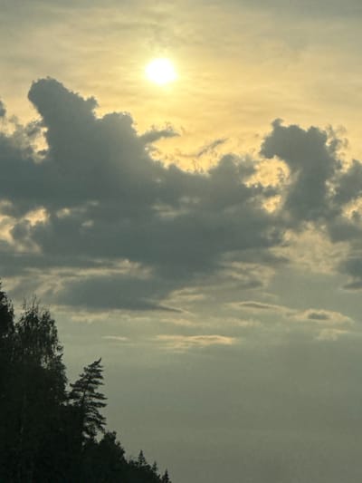 The image depicts a sky filled with clouds and the sun, viewed from a forested area in Khlyupinskogo Lesn-Va, Russia. The sun, a bright, white orb, is positioned in the upper left quadrant of the frame, casting a warm, yellow glow through the scattered clouds. The clouds themselves vary in shade from dark gray to lighter tones, with some areas illuminated by the sun. In the lower left portion of the image, the silhouettes of trees and a forest edge are visible, suggesting a natural, outdoor setting. The overall impression is one of a serene, possibly late afternoon or early morning sky. There are no people or discernible objects in the foreground or mid-ground.