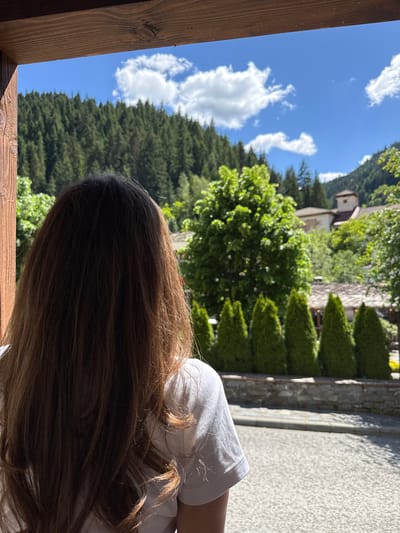A young person with long, brown hair, viewed from behind, stands looking out at a scenic view of a mountainside covered in dense green pine trees under a bright blue sky with scattered white clouds. In the mid-ground, several conical evergreen trees are planted in a row, backed by a stone wall and some traditional Bulgarian houses with red tile roofs, one featuring a prominent tower. The scene is framed by a wooden overhang in the foreground, suggesting the person is looking out from a covered porch or balcony. The overall impression is one of peaceful contemplation in a rural mountain village setting, likely Shiroka Laka, Bulgaria.
