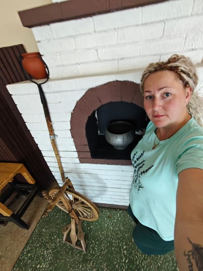 In an interior setting, a Caucasian woman with blonde hair styled in dreadlocks stands to the right of a traditional Russian stove, which is constructed of white brick with a brown, arched opening. A clay pot rests on a long, wooden tool beside the stove. To the left of the stove, a wooden spinning wheel is positioned on a speckled green floor. The woman is wearing a light blue t-shirt with a black floral design and dark green trousers. She has a tattoo of a feather and lettering on her right forearm. The environment suggests a rural or historical dwelling in Cheryomushki, Russia.