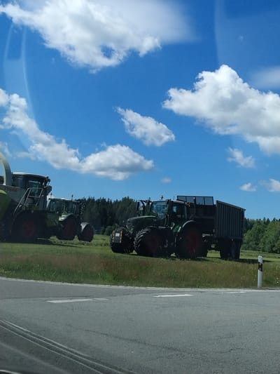 The image depicts an outdoor scene on a sunny day, likely in a rural area near Hinterschmiding, Germany. In the foreground, a grey asphalt road is visible with white lane markings. Beyond the road, a grassy field stretches out, dotted with some wildflowers. Several agricultural machines are parked on the field.

On the left side of the image, a large, green and grey CLAAS forage harvester is positioned. To its right, two green Fendt tractors are parked. The tractor on the left is smaller than the one on the right. The Fendt tractor on the right is connected to a dark grey, large-capacity trailer. In the background, a dense line of dark green trees forms a forest. The sky is bright blue with scattered white, fluffy clouds, suggesting fair weather. No people are visible in the frame. The overall impression is one of agricultural machinery at rest, likely during a break in field work.
