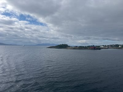 The image depicts a view of the Vestfjorden from Lødingen, Norway. The water is a deep, dark blue, with small waves indicating a moderate chop. In the distance, a chain of islands and mainland shores stretches across the horizon under a cloudy sky. On one of these islands, a small settlement is visible, characterized by red-roofed buildings, including a prominent red boathouse with a pier. A lighthouse stands on the highest point of this island. Further to the left, a white ferry or ship is navigating the waters. The overall atmosphere is overcast and somewhat muted, typical of the Norwegian coast.