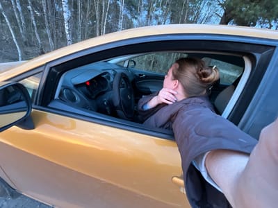 A medium shot captures a light-skinned woman in the driver's seat of a yellow car. She has light brown hair styled in a bun and is wearing a dark, puffy jacket with a white sleeve visible at the wrist. Her left hand is near her face. The car's interior, including the dashboard and steering wheel, is visible. Outside the car window, a blurred background of trees suggests a wooded area. The angle suggests the photo was taken from outside the car, with the photographer's arm and hand partially visible in the frame. The location is Voronino, Russia.