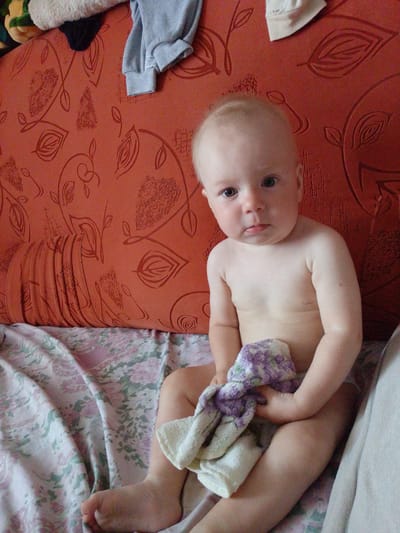 The media file shows a close-up, high-angle shot of a baby sitting on a patterned surface, likely a bed or sofa. The baby is facing the camera with a neutral expression. They are holding a folded towel or blanket with a purple and white floral pattern. The baby is nude from the waist up, with their legs obscured by the towel and the patterned surface. The background behind the baby is a textured, orange surface with a repeating leaf pattern. On the orange surface, there are also some pieces of clothing, including a light blue garment and a white cap. The lighting appears to be natural, suggesting it might be daytime. The setting seems to be indoors, possibly a bedroom or living room. The location context is Birsk, Russia. There is no visible text in the image.