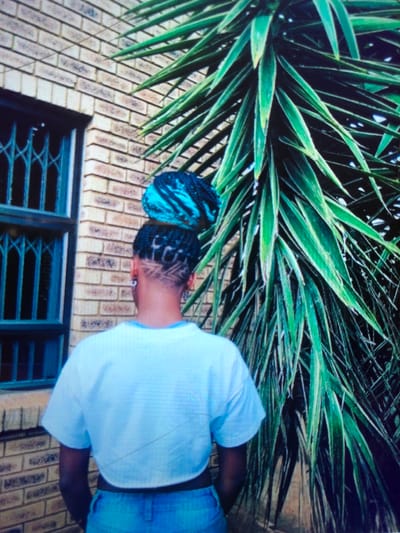 An individual stands with their back to the camera in Soweto, South Africa. The subject has dark skin and a distinctive hairstyle featuring a shaved nape with a precise geometric pattern, braids above the nape arranged to form letter-like shapes, and a voluminous top bun composed of blue and black braids. A silver stud earring is visible in the left ear. The individual wears a light blue, short-sleeved crop top and light blue denim jeans. The background on the left consists of a light brown brick wall with a dark-framed window secured by a metal security grille. To the right, a large plant with long, sword-shaped green leaves extends vertically, partially obscuring the background. The scene is illuminated by natural daylight.