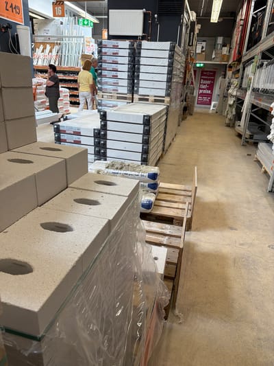 This image appears to be taken inside a hardware store, likely in Gütersloh, Germany, given the German language signs. The store is well-stocked, with shelves filled with various construction and hardware items, including what seems to be piping, insulation materials, and stacks of building blocks wrapped in plastic. At the foreground, there's a prominently displayed stack of concrete blocks with circular holes.
Several people are present in the middle ground, browsing the merchandise. There are two women wearing yellow and green tops respectively, and one person with a gray T-shirt, inspecting the products.
Signs in the store advertise "Dauer-Tief-Preise" (long-term low prices), indicating special offers. The image's composition, with the stacks of materials and people moving through the aisles, suggests a typical retail environment for construction and home improvement goods.