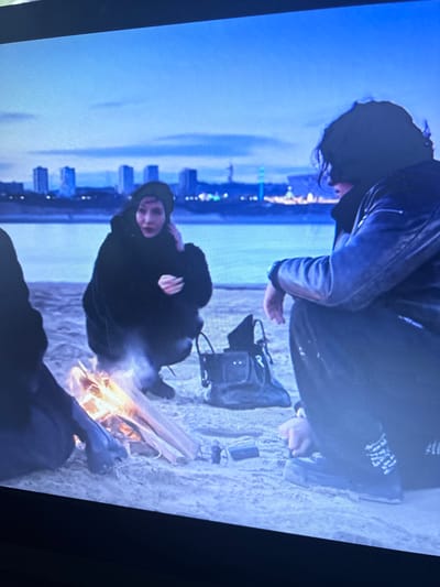 The image depicts two individuals on a beach, sitting near a small bonfire. In the background is the skyline of a city, believed to be Cheongju-si, South Korea. One person, facing forward, is hooded and wrapped in dark clothing. The second person, clad in a leather jacket, is crouched down, with their hand extended toward an object on the ground next to a black bag. The beach sand appears light, and there are a few other scattered objects near the fire. The sky is a gradient of blue.