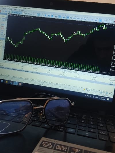 The image depicts an indoor scene in Yangiabad, Uzbekistan, featuring a close-up view of a black laptop computer. The laptop's screen displays a financial trading platform, specifically a candlestick chart with green and white candles against a dark background, showing an upward market trend. Various numerical data, timestamps, and interface elements, some in Cyrillic script, are visible on the screen, including a system time of "14:47 18.01.2038". A pair of modern eyeglasses with a light metallic frame and dark, reflective, polygonal lenses rests on the laptop's black keyboard. In the bottom right foreground, a portion of a white rectangular object, identified by text as "Aspire E 15" and "USB 3.1" along with a Wi-Fi icon, is partially visible. No individuals are present in the frame.