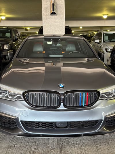 The image shows the front of a dark gray BMW M car parked in an indoor setting, likely a garage or parking structure. The car is positioned centrally in the frame, with its distinctive grille, headlights, and hood clearly visible. The grille features the iconic BMW emblem and three colored stripes – red, light blue, and dark blue – indicating it's a performance model. The car's headlights are detailed and modern, and the hood has a reflective sheen, highlighting the car's sleek design.

In the background, other vehicles are partially visible, suggesting a parking lot or a car dealership. The setting is dimly lit, with warm yellow lights illuminating the walls and parts of the vehicles. The walls appear to be concrete or a similar industrial material. To the left of the car, a concrete pillar with a metallic fixture is visible, adding to the utilitarian feel of the location. The overall impression is one of a well-maintained, high-performance vehicle in a secure, indoor environment. There is no visible text in the image. The time of day and weather are not discernible from the image.