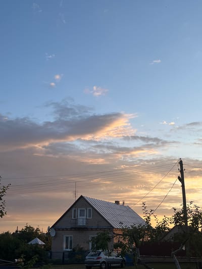 The image depicts a rural scene in the Sarootski Sel'ski Savet, Belarus, during what appears to be sunset. A two-story brick house with a silver metal roof is prominently featured, with a balcony on the upper level. Adjacent to the house, a white car is parked. Power lines traverse the sky, supported by a wooden utility pole. Lush green trees and foliage surround the property. The sky is a blend of blue, with scattered clouds tinged with warm hues of orange and pink, indicating the setting sun. The overall impression is one of a peaceful, rural dwelling at dusk.