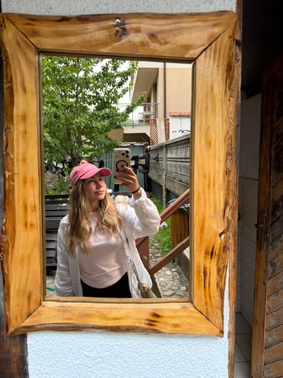The image is a mirror selfie taken by a young woman with blonde hair and a pink baseball cap. She is wearing a white shirt and a pink top. She is holding a smartphone in her right hand, using it to take the photo. The mirror is framed by a rustic wooden border with a natural, unfinished edge.
The background reflected in the mirror shows a residential area with buildings and lush green trees. The architecture suggests a European setting, consistent with the location context of Konjic, Bosnia and Herzegovina. There are glimpses of stone walls and outdoor furniture, hinting at a courtyard or patio.
The lighting suggests it is daytime, likely a bright day given the clear reflections. The overall mood appears casual and relaxed. There is no visible text in the image, other than what might be on the woman's phone case or the branding of the phone itself, which is not discernible.