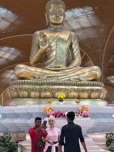 The image depicts a large, golden Buddha statue seated on a tiered platform in a well-lit indoor space. In the foreground, a man in a red shirt is taking a selfie with a woman in a pink shirt. Behind them, another person in a black shirt is also visible. The platform is decorated with floral arrangements and has a series of layered steps. A sign with text in Khmer and English is also present to the left of the people. The background features a distinctive patterned ceiling with circular lights. The presence of the sign in Khmer suggests the location is in Cambodia. Given the context of the large Buddha statue and the surrounding environment, this appears to be a significant religious or cultural site. The city of Kandal Stueng is known for its pagodas and cultural heritage, making it a plausible location for such a monument.