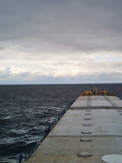 The image shows a ship's deck on a cloudy day. The deck is gray and textured, with a line of circular hatches running down the middle. Blue railings line the sides of the deck. Beyond the deck, the dark water of a sea or large lake is visible, with white-capped waves. In the distance, a landmass is visible on the horizon. The sky is overcast with gray clouds, suggesting stormy weather. The location is likely near Prislon', Russia, based on the reference to a city with that name, but no specific elements confirm it. No people are visible in the image. The ship appears to be moving, given the waves.