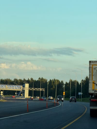The image captures a view of a highway in Egor'evo, Russia, under a pale blue sky with scattered clouds. A red car is visible on the road approaching a pedestrian overpass. Several other vehicles, including SUVs and cars, are driving on the highway. Traffic cones and streetlights line the road. A large truck is partially visible on the right side of the image. The highway is surrounded by dense green trees.