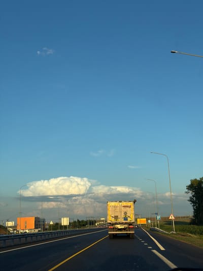 An eye-level, forward-facing shot captures a section of a highway in Egor'evo, Russia, under a bright blue sky with scattered clouds. The road, paved with asphalt, features a yellow double line marking the center and white dashed lines on the right side. A large, white semi-trailer truck is driving away from the viewer in the right lane. The truck has "MEZA LIGHTS" printed on its rear. Streetlights line the right side of the highway, interspersed with various road signs, including a triangular warning sign with a red border and a white exclamation mark. On the left side of the road, an orange building with dark windows stands behind a metal guardrail. In the distance, more streetlights and road signs are visible, along with a line of trees to the far right. The overall scene depicts a typical highway environment during daylight hours.