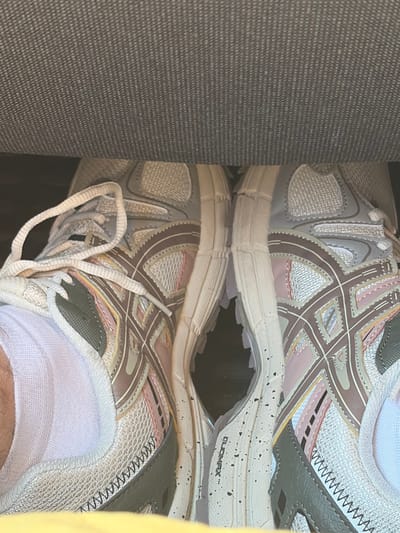 A close-up, top-down view shows a pair of athletic shoes, likely running shoes, resting on a textured, dark gray fabric surface, possibly part of a seat or upholstery. The shoes are predominantly white with brown, pink, and gray accents, featuring mesh panels and crisscrossing decorative stitching. One shoe on the left is slightly angled, with its white laces loosely tied, revealing a white sock on the wearer's foot. The other shoe on the right is positioned similarly. The overall lighting suggests an indoor environment. The image appears to have been taken in the Settlement of sanatorium "Podmoskovye", Russia.