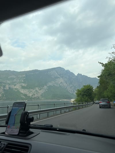 The image, taken from inside a vehicle, shows a view of the landscape near Mandello del Lario, Italy. The foreground includes a car dashboard with a phone mounted displaying a navigation app. Through the windshield, a winding road is visible, flanked by trees and a guardrail. In the distance, a large, rugged mountain dominates the scene. The sky is overcast with a gray, cloudy sky.