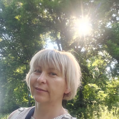 The media file is a selfie taken outdoors, likely in a park or wooded area, given the abundance of trees and dappled sunlight. The time of day appears to be late afternoon or early evening, as indicated by the warm, low-angled sunbeams breaking through the foliage. The weather seems pleasant, with clear skies allowing for bright sunlight.

The main subject is a woman with blonde, shoulder-length hair. She is smiling slightly, and her face is partially turned towards the camera. She is wearing a striped top with a strap visible over her shoulder. Her expression suggests a relaxed and content mood.

The background is dominated by lush green trees, with the sun creating a strong lens flare effect as it shines through the leaves. The scene is vibrant and natural, evoking a sense of peace and tranquility. There are no visible text elements in the image. The context of Voronkiv, Ukraine, suggests this might be a personal snapshot taken during leisure time in a local park or natural area.