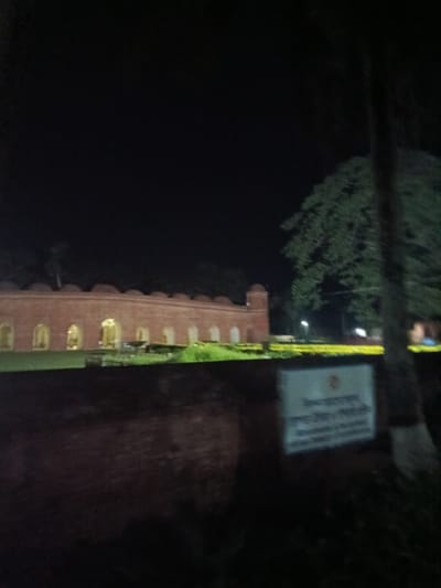 The image captures a night scene of the Khan Jahan Ali's tomb complex in Bagerhat, Bangladesh. A long, arched brick structure, likely part of the historical site, is illuminated by interior lights, casting a warm glow through its arched openings. The foreground features a dark brick wall, with a white sign displaying Bengali text positioned on its surface. A large tree with dense foliage is visible on the right side of the frame, its dark silhouette contrasting with the lighter background. The overall ambiance is dark and quiet, characteristic of a historical monument at night.