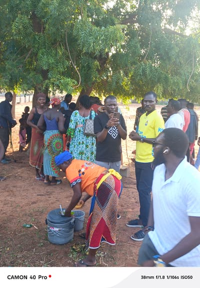A medium shot shows a group of people, numbering about fifteen, gathered in Gidan Kogi, Nigeria. The setting is outdoors, under the shade of a large tree with full, green foliage.

In the foreground, a woman wearing an orange shirt, blue headwrap, and a multi-colored patterned skirt bends over two gray buckets and a large gray food container. She appears to be serving or retrieving something from the container.

Around her, the group includes men and women of varying ages, some standing and talking, others observing the woman with the buckets. A man dressed in a black shirt with a geometric pattern is holding a smartphone, appearing to take a picture. Another man in a yellow soccer jersey stands nearby. Other people are further in the background. The ground is red dirt.