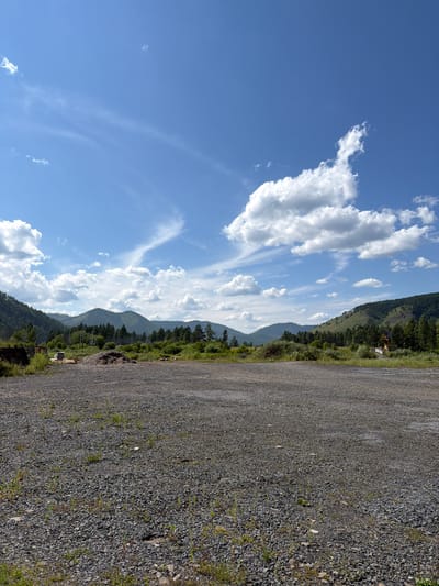 An outdoor, eye-level, vertical shot captures a landscape under a bright blue sky with scattered white clouds. The foreground is dominated by a rough, grey gravel surface with sparse green vegetation poking through. In the midground, a line of dark green coniferous trees stretches across the scene, with lighter green foliage and some bare patches visible. Beyond the trees, rolling, verdant hills rise, their slopes dotted with more trees. The sky features wispy cirrus clouds and larger cumulus formations, particularly on the right side of the frame. There are no people or animals visible in the image. The scene appears to be an undeveloped area or a clearing in a natural landscape, possibly on the outskirts of Bol'shoe Goloustnoe, Russia, given the presence of mountainous terrain and dense forestation characteristic of the region.