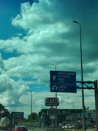 The image shows a street scene in Ukraine, likely on Peremohy Street, under a partly cloudy sky with a teal tint. A large blue directional signpost indicates routes to Zhytomyr (straight ahead) and Chop (straight ahead), and Kyiv (turn left). Below this, a sign for "ATB MARKET" is visible, along with its operating hours "00-24" and designations for "ВЛАСНА ВИПІЧКА" (homemade baked goods) and "ВЛАСНА КАВ'ЯРНЯ" (own coffee shop). To the left, a truck with "БЕТОН" (concrete) written on its side is parked, and a red car is in the foreground. A tall street lamp stands prominently on the right side of the image. In the background, a gas station and several other vehicles are present, along with a few pedestrians.