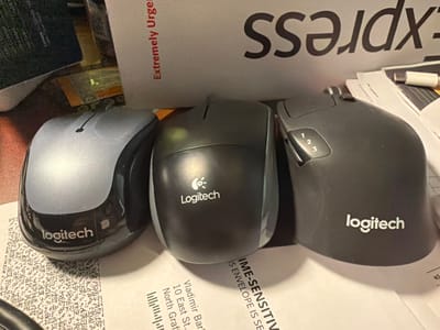 Three computer mice are positioned on top of various documents. The mouse on the left is silver and black with a "logitech" logo. The middle mouse is dark gray, also with a "Logitech" logo and a small leaf-like symbol. The mouse on the right is dark gray and features numbered buttons "1", "2", and "3" as well as a "logitech" logo. The documents beneath the mice appear to be shipping labels and other paperwork, with text visible that includes "Extremely Urgent", "IS ENVELOPE IS SE", and "North Graff". The background shows part of a box or package with the letters "ss" and "rdx" visible, suggesting it may be related to a shipping company. The setting appears to be an indoor environment, possibly a desk or workspace. No people are visible in the image. There is no information within the image to indicate the specific location within the city of Grafton, United States of America.