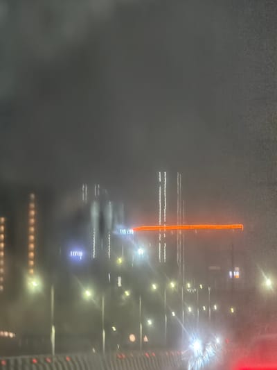 The image depicts a blurry, nighttime scene in what appears to be a city. Dominating the background are tall, illuminated buildings, some with strings of lights resembling windows or architectural features. In the foreground, streetlights cast a hazy glow, and a prominent horizontal orange light streak suggests motion, possibly from a vehicle or a lit sign. The overall atmosphere is obscured by what appears to be fog or heavy rain, creating a diffused and somewhat ethereal quality. The presence of a road barrier in the lower portion of the image indicates a transportation route, likely a highway or major road. The location is identified as Manchirevula, India. No people are clearly discernible in the image due to the blurriness and low light conditions.