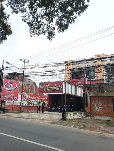 A street view features the "Sambal Ndower" restaurant, its signage prominently displayed alongside a banner welcoming patrons and advertising a price of 8,000.00. Several motorcycles are parked near the establishment's entrance, hinting at its popularity. Overhead, a dense network of power lines intersects the sky. Graffiti adorns a nearby building, while sparse foliage frames the top of the image. The overcast sky casts a muted light over the scene, captured from a slightly low angle along the roadside.