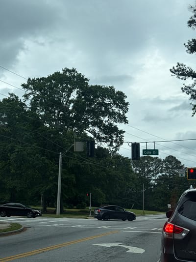 The image depicts a street scene in Mountain Park, United States of America. The view is from a vehicle at a traffic light on the corner of Cole Dr and Highway 78. The traffic light is red and there are trees around the intersection. There is a Nissan Pathfinder in front of the vehicle from which the picture was taken. There is another car traveling in the opposite direction turning on Cole Dr. The sky is overcast.