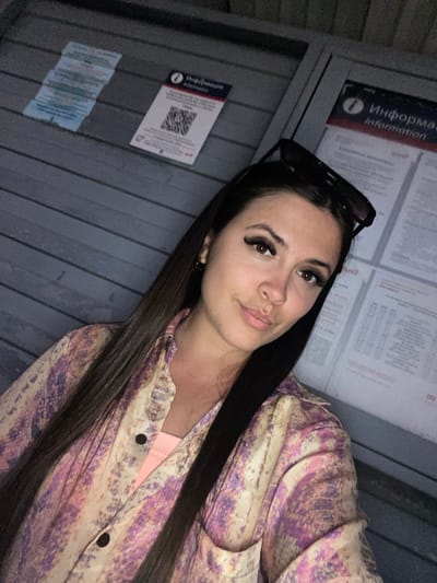 A young woman with long, dark brown hair, wearing a pink and purple patterned button-down shirt over a peach-colored tank top, poses for a selfie outdoors at dusk. She has false eyelashes and a neutral expression, with sunglasses perched on top of her head. Behind her, a grey, horizontally slatted wall serves as the backdrop, with two informational posters affixed to it. One poster displays a QR code and Russian text that translates to "Information." The other poster also contains Russian text. The lighting suggests an urban environment, possibly a bus stop or public information kiosk in Samur, Russia.