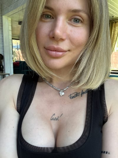 A close-up selfie captures a young woman with fair skin and shoulder-length, light blonde hair. Her eyes are a light grey-blue or green, and her lips are full with a natural, glossy finish. She sports a serene and slightly smiling expression. She is wearing a black ribbed top with sheer mesh detailing along the neckline and straps, revealing her upper chest. A delicate silver-toned necklace, featuring a prominent heart-shaped pendant, is worn around her neck.

Two tattoos are visible on her chest area. On her left collarbone, a script tattoo reads "todo feito por," which is Portuguese/Spanish for "all made by" or "everything done by." On her right chest, a Cyrillic script tattoo clearly spells out "Сияй!", translating to "Shine!" in Russian.

The photograph is well-lit by natural light, suggesting it was taken during the daytime. The background is softly blurred but appears to be an indoor setting, with hints of reflective surfaces and light-colored walls or tiles, possibly suggesting a bathroom or a bright room with windows. The presence of the Cyrillic tattoo aligns with the provided location context of Ozernoe, Russia. The woman's pose is relaxed, and her gaze is directed towards the camera.
