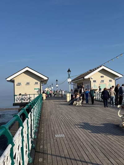 The image depicts Penarth Pier in Penarth, United Kingdom, on a clear, sunny day. The pier features a wide wooden plank deck extending from the foreground into the background, where numerous pedestrians are present.
On the left side of the pier, a decorative green and white metal railing separates the walkway from the calm water and exposed mudflats below. Towards the mid-left, a light yellow single-story building with a dark sloped roof and white trim is situated over the water, supported by the pier structure. It has small, rectangular windows on its side and two individuals are visible seated at a table on a small balcony beneath it.
Further along the pier to the right, another similar light yellow building with a dark roof and white trim stands on the deck. This building features larger windows and appears to house commercial activity, with several people standing near its entrance or displays. A string of white globe lights is strung diagonally from the roof of this building towards the upper center of the image.
Multiple black ornate lamp posts are spaced along the pier. Various individuals and small groups are seen walking, standing, and interacting. In the foreground-right, two individuals are seated on a white decorative bench. Shadows are cast across the wooden deck, indicating strong sunlight from the left. The sky is clear and blue, without clouds.