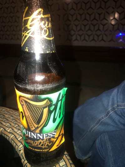 A close-up shot shows a bottle of Guinness Smooth stout lying on its side. The label features the iconic Guinness harp in gold, transitioning into a green and yellow ombre effect. "Guinness Smooth" is prominently displayed on the label. The bottle is brown and slightly wet, appearing to have condensation. Part of a pair of blue jeans is visible in the upper right corner, suggesting someone is seated nearby. A patterned surface is seen in the upper left, possibly a tabletop or wall.
