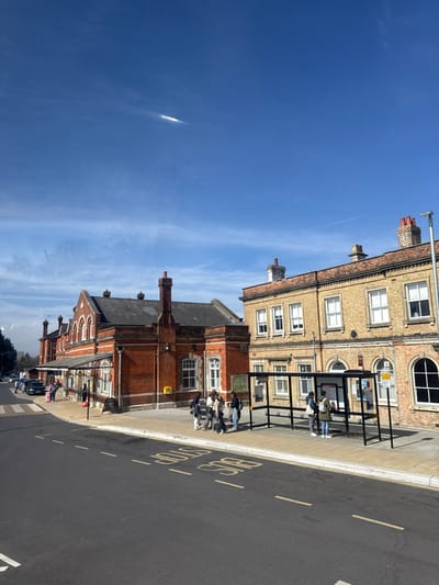 The image depicts an exterior street scene on a clear, sunny day in Salisbury, United Kingdom, specifically showing the frontage of Salisbury railway station and an adjacent building.
In the foreground, a dark asphalt road surface with "BUS STOP" painted in white text (reversed from this perspective) and double white dashed lines is visible. A grey paved sidewalk separates the road from the buildings.
On the sidewalk, a modern bus shelter with a black metal frame and transparent panels stands prominent. Inside the shelter, three individuals are present, observing information displays. To the left of the bus shelter, four more individuals are gathered on the pavement, appearing to be conversing.
The dominant structure on the left is the Salisbury railway station, a two-story building constructed of red brick with dark grey roof tiles and multiple brick chimneys. It features arched windows, decorative brickwork, and a dark-colored awning extending over a portion of the sidewalk. A yellow utility box or post box is affixed to its facade. Further down the street to the left, several vehicles are parked, and one person with a pink suitcase is visible near a car.
Immediately to the right of the station is another two-story building, constructed of lighter yellow brick with darker brick details and chimneys. This building also features multiple windows, some arched on the ground floor, and a decorative cornice line.
The sky overhead is clear and blue, with minimal wispy clouds and a distinct, bright white contrail or atmospheric phenomenon visible high in the upper-center of the frame.