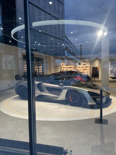 The image depicts a car dealership viewed through a window, likely in Glattbrugg, Switzerland based on visual cues and potential location context. Three cars are visible, one in silver, one in black, and one in red and black, all positioned in a row within the dealership. A circular, illuminated platform showcases the foremost silver car. In the background, a shelving unit displays various items. The window through which the scene is viewed reflects the exterior environment, including a building with a distinctive arched light fixture. No people are visible.