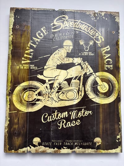 This image depicts a vertically oriented, rectangular wooden sign, consisting of multiple dark-stained wood planks joined together, displaying a vintage-style design in metallic gold. The sign exhibits deliberate distressing, including chipped edges, visible wood grain, knots, and areas where the gold design appears faded or scratched.

The central subject is a stylized illustration of a motorcycle racer on a vintage motorcycle, depicted in profile, moving from left to right. The racer wears a helmet and a full racing suit, leaning forward over the handlebars. The motorcycle features large spoked wheels, an exposed engine block, and a solo seat. Horizontal lines behind the rider and motorcycle suggest speed.

Various text elements are integrated into the design. Vertically along the left edge, "VINTAGE" is printed. Vertically along the right edge, "RACE" is printed. Arched across the top center, the word "Speedmasters" is prominent, with "DESIGNS CALIFORNIA" directly below it. Below "VINTAGE" on the left, "ALL PRODUCT MADE OF THE BEST SERIES" is visible. Near the top-right, above the motorcycle engine, a graphic of two pistons and connecting rods is accompanied by the text "OF THE BEST SERIES." At the bottom center, in a large script font, "Custom Motor Race" is displayed. Below this, "National Championships" is printed, followed by "STATE FAIR TRACK MILWAUKEE." Small, stylized helmet graphics are positioned in the bottom corners of the sign. Two screws are visible near the top left and top right corners, securing the sign.

The overall scene is a decorative object designed to evoke a rustic, retro aesthetic associated with motorcycle racing. The image is located in the city of West Felton, United Kingdom.