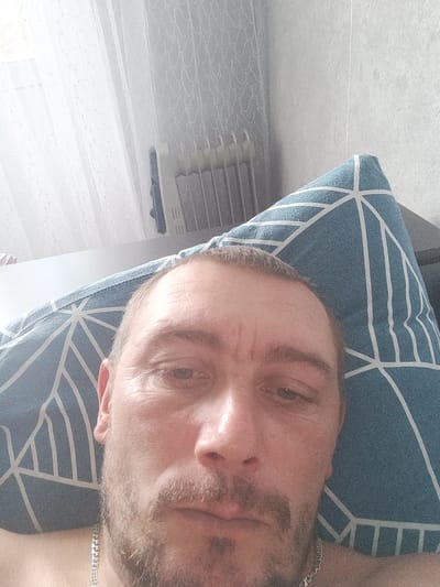 The image shows a close-up, downward-angle selfie of a man resting on a pillow. The man has fair skin, short brown hair, and a beard. He is wearing a silver chain necklace. The pillow is blue with a white geometric pattern. In the background, a white sheer curtain is visible, and to the right of the curtain, a radiator with vertical fins can be seen against a light gray textured wall. The scene appears to be indoors, likely in a bedroom. The lighting suggests it could be daytime, with natural light coming from the left. The image is pixelated and lacks sharp detail. The overall impression is of a casual, personal moment captured by the subject. There is no information in the image to suggest the location is Novosemeykino, Russia.