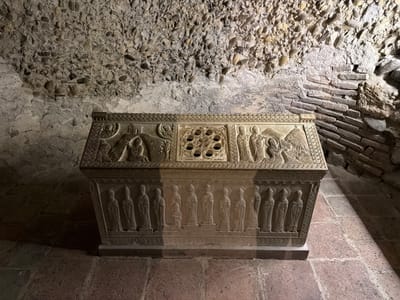 An ornate, stone sarcophagus with carved reliefs sits in a dimly lit, ancient chamber. The sarcophagus is rectangular with a slightly sloped lid, covered in intricate carvings depicting religious figures and scenes. The lid features a central decorative element with circular openings and various figures arranged around it, including what appears to be a Nativity scene on the left and other figures on the right. The front of the sarcophagus displays a row of standing figures, each with halos, and a small kneeling figure in the center. The stone is a light, weathered beige. The chamber walls are constructed from rough stone and brickwork, with a textured ceiling of pebbles and mortar, suggesting an underground or historical setting. The floor is paved with reddish-brown stone tiles. The lighting casts long shadows, emphasizing the texture and depth of the carvings and the rough surfaces of the walls.